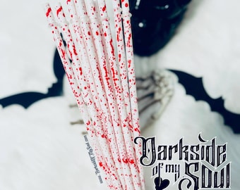 May include: A bundle of white plastic drinking straws with red splatters, resembling blood, on them. The straws are held in a hand with black nail polish. The background features a black skull and a bat silhouette.