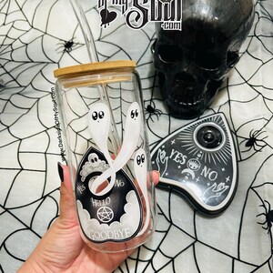 Pu&ograve; includere: Un bicchiere di vetro trasparente con un coperchio in bamb&ugrave; e una cannuccia. Il bicchiere ha un design bianco di una tavola Ouija con fantasmi e le parole "YES", "NO", "HELLO" e "GOODBYE".