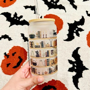 May include: A frosted glass tumbler with a bamboo lid and a straw. The tumbler features a Halloween-themed design with a bookshelf filled with books, candles, pumpkins, a black cat, and a ghost. The text "www.DarksideOfMySoul.com" is printed on the side of the tumbler.