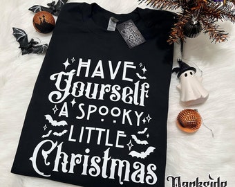 May include: Black t-shirt with white text that reads "Have Yourself A Spooky Little Christmas". The text is in a vintage font and is surrounded by small bats and stars. The shirt is on a white surface.