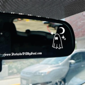 May include: A rearview mirror with a white ghost sticker on it. The ghost has two eyes and is surrounded by stars and a crescent moon. The text "www.DarksideOfMySoul.com" is visible at the bottom of the mirror.