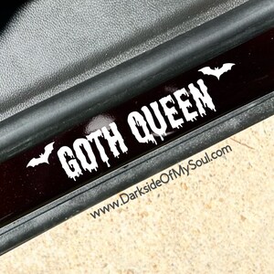 May include: A black and white sticker with the text "GOTH QUEEN" in dripping letters and two bats on either side. The sticker is on a black surface with a website address below the text: www.DarksideOfMySoul.com