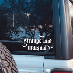 May include: White vinyl decal with the text "strange and unusual" in a gothic font with three bat silhouettes. The decal is on the back window of a silver car.