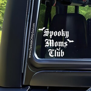 May include: White vinyl decal with the text "Spooky Moms Club" in a gothic font. The decal is on the back window of a black car. There are three bat silhouettes above and below the text. The text "www.DarksideOfMySoul.com" is on the side of the car window.