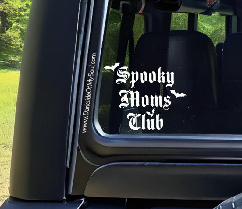 May include: White vinyl decal with the text "Spooky Moms Club" in a gothic font. The decal is on the back window of a black car. There are three bat silhouettes above and below the text. The text "www.DarksideOfMySoul.com" is on the side of the car window.