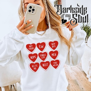 May include: White sweatshirt with red heart-shaped candy graphics. Each heart has a different Valentine's Day themed message, including "Not Yours", "EW", "Boy Bye", "Nope", "Single Life", "Meh", "Bite Me", "Next", and "You Wish".