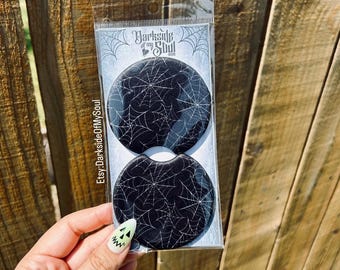 May include: Two circular black car cup holder coasters featuring a white spiderweb pattern. The coasters are packaged in a clear plastic sleeve with a header card displaying the text Darkside of my Soul and Etsy:DarksideOfMySoul.