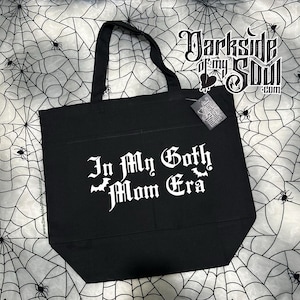 Puede incluir: Bolsa de tela negra con texto blanco que dice "In My Goth Mom Era" con un gr&aacute;fico de murci&eacute;lago. La bolsa tiene un bolsillo en la parte delantera y est&aacute; hecha de lona.