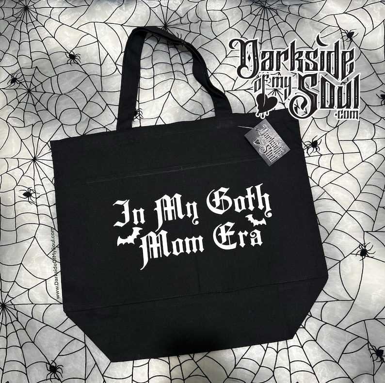 Puede incluir: Bolsa de tela negra con texto blanco que dice "In My Goth Mom Era" con un gr&aacute;fico de murci&eacute;lago. La bolsa tiene un bolsillo en la parte delantera y est&aacute; hecha de lona.
