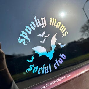 May include: A holographic sticker with the text "Spooky Moms Social Club" in a gothic font. The sticker features a large bat with smaller bats flying around it. The text "www.DarksideOfMySoul.com" is at the bottom of the sticker.