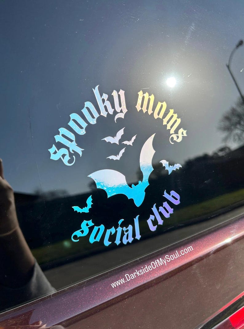 May include: A holographic sticker with the text "Spooky Moms Social Club" in a gothic font. The sticker features a large bat with smaller bats flying around it. The text "www.DarksideOfMySoul.com" is at the bottom of the sticker.