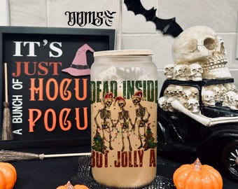 May include: A Halloween-themed tumbler with a festive design. The glass features a skeleton graphic with the text "DEAD INSIDE BUT JOLLY AF". The scene includes pumpkins, a sign that says "IT'S JUST HOCUS POCUS", and decorative bats.