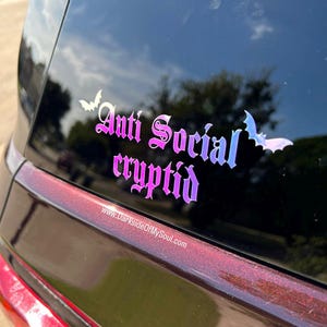 May include: A black car with a rear window sticker that reads "Anti Social Cryptid" in a gothic font with a purple and blue iridescent finish. The sticker also features two bat silhouettes.