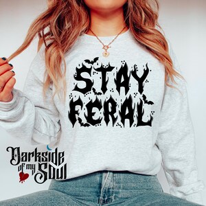 May include: A grey sweatshirt with the text "STAY FERAL" in black, styled with flames and bats.