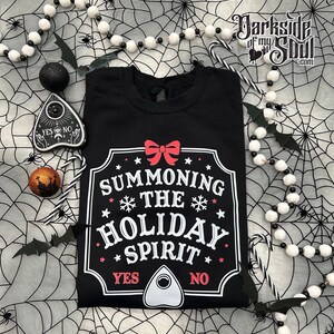 Op de afbeelding: Zwart sweatshirt met de tekst "SUMMONING THE HOLIDAY SPIRIT" in wit en rood. Het ontwerp bevat een rode strik, sneeuwvlokken en sterren. Halloween-decoraties omringen het sweatshirt.