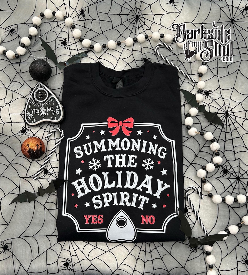 Op de afbeelding: Zwart sweatshirt met de tekst "SUMMONING THE HOLIDAY SPIRIT" in wit en rood. Het ontwerp bevat een rode strik, sneeuwvlokken en sterren. Halloween-decoraties omringen het sweatshirt.