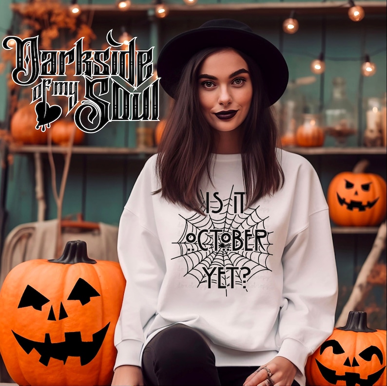 K&ouml;nnte beinhalten: Ein wei&szlig;es Sweatshirt mit einem schwarzen Spinnennetz-Design und dem Text "Is it October yet?" auf der Vorderseite. Das Model tr&auml;gt einen schwarzen Hut und steht vor einer mit K&uuml;rbissen dekorierten Wand.