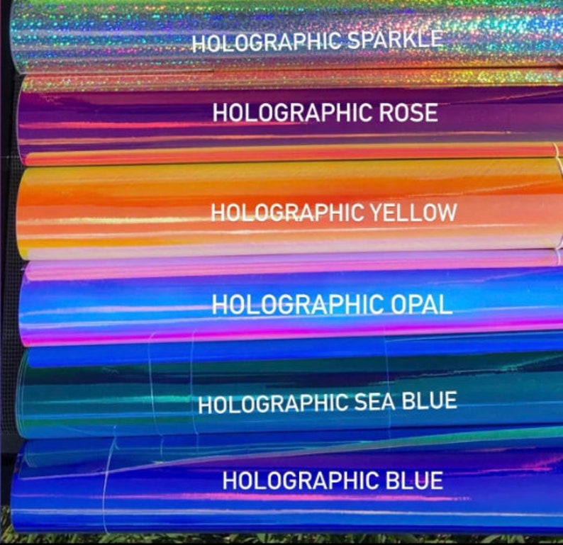 May include: A stack of six rolls of holographic vinyl in various colors, including sparkle, rose, yellow, opal, sea blue, and blue. Each roll has a label with the color name and the word "Holographic".