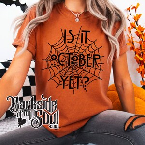K&ouml;nnte beinhalten: Eine Frau tr&auml;gt ein orangefarbenes T-Shirt mit einem schwarzen Spinnennetz-Motiv und dem Text "Is it October Yet?" darauf gedruckt. Sie tr&auml;gt graue Jeans und wei&szlig;e Schuhe.