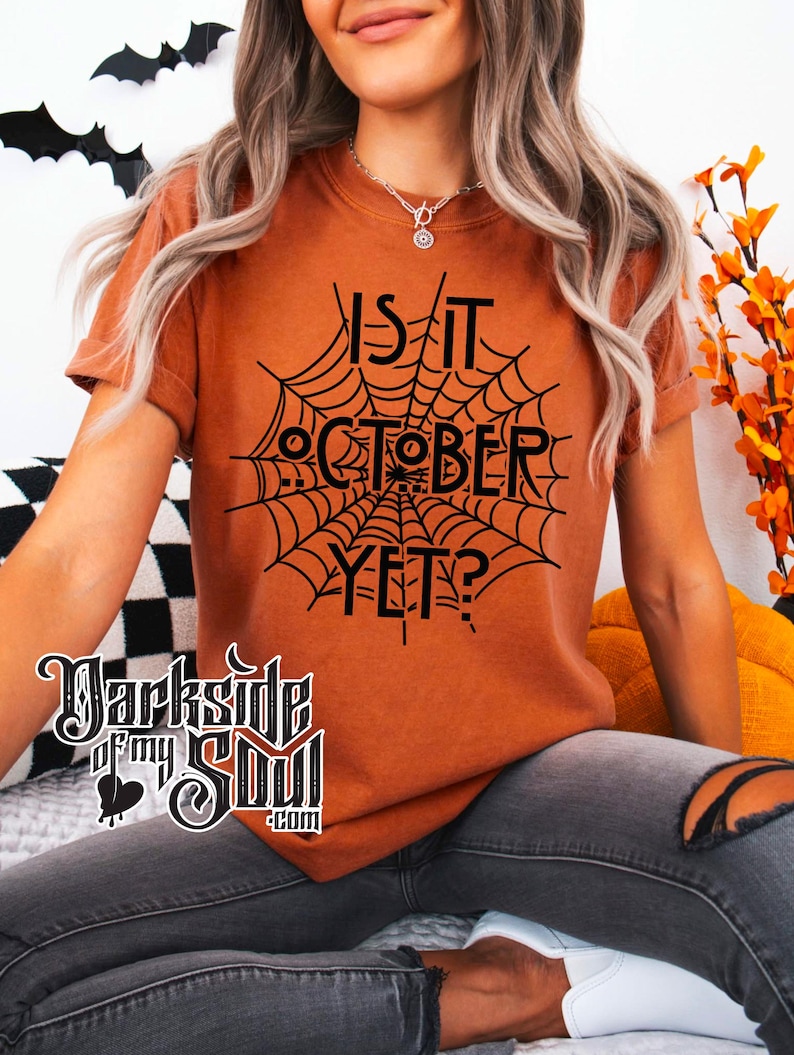 K&ouml;nnte beinhalten: Eine Frau tr&auml;gt ein orangefarbenes T-Shirt mit einem schwarzen Spinnennetz-Motiv und dem Text "Is it October Yet?" darauf gedruckt. Sie tr&auml;gt graue Jeans und wei&szlig;e Schuhe.