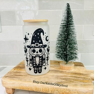 May include: A frosted glass can-shaped tumbler with a bamboo lid, featuring a black graphic of a skeleton nutcracker wearing a witch's hat. A small green Christmas tree sits next to the tumbler on a wooden serving board. The board has the text "Etsy: DarksideOfMySoul".