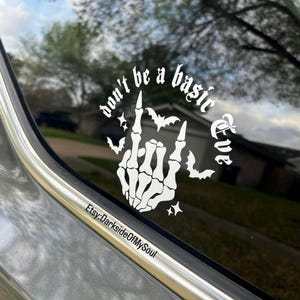 May include: White vinyl decal on a car window. The decal features the phrase "don't be a basic Eve" with a skeleton hand making a rock and roll gesture, bats, and stars. The Etsy shop name "DarksideOfMySoul" is also included.