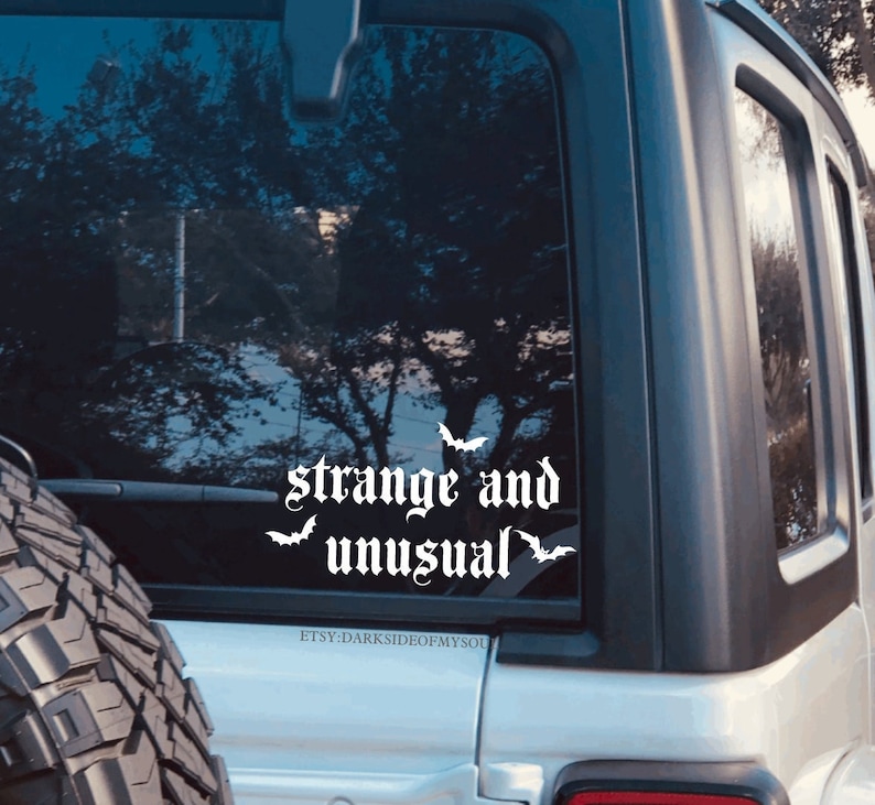 May include: White vinyl decal with the text "strange and unusual" in a gothic font with three bat silhouettes. The decal is on the back window of a silver car.