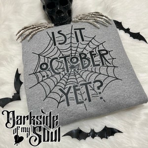 K&ouml;nnte beinhalten: Ein graues Sweatshirt mit einem schwarzen Spinnennetz-Design und dem Text "Is it October yet?" darauf gedruckt.