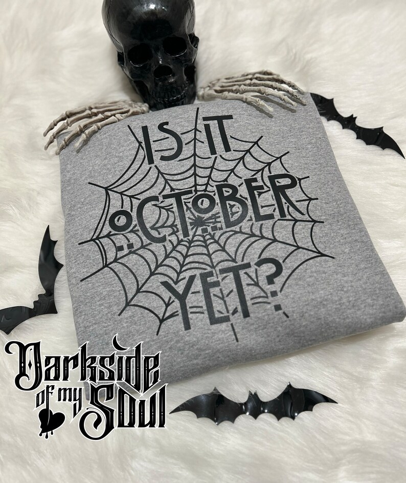 K&ouml;nnte beinhalten: Ein graues Sweatshirt mit einem schwarzen Spinnennetz-Design und dem Text "Is it October yet?" darauf gedruckt.