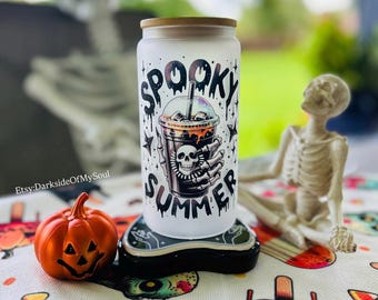 May include: A frosted glass tumbler with a bamboo lid featuring a graphic of a skeleton hand holding an iced coffee cup with a skull design. The text "SPOOKY SUMMER" is printed in a dripping black font around the central illustration.
