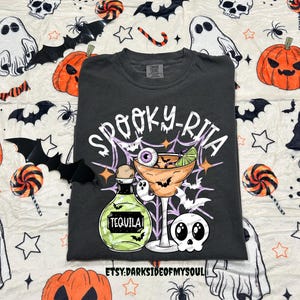 May include: Dark gray t-shirt with a Halloween-themed graphic. The design features a margarita glass with an eyeball, a tequila bottle, bats, and the text "Spooky-Rita". The shirt is surrounded by Halloween-themed decorations.