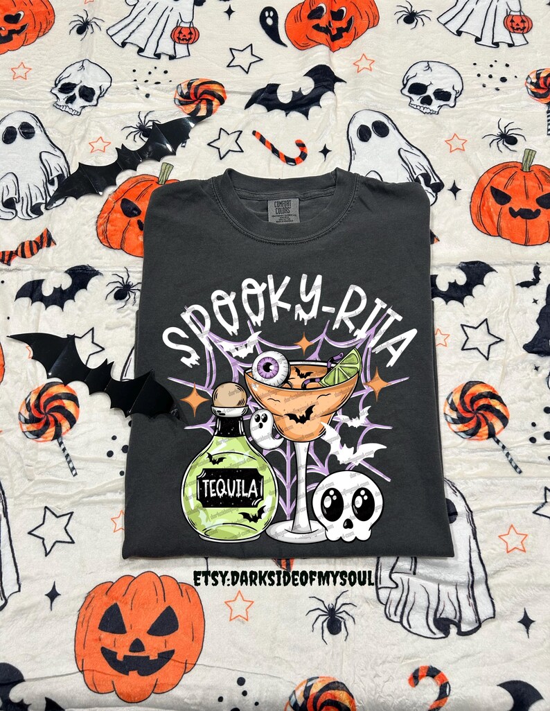 May include: Dark gray t-shirt with a Halloween-themed graphic. The design features a margarita glass with an eyeball, a tequila bottle, bats, and the text "Spooky-Rita". The shirt is surrounded by Halloween-themed decorations.