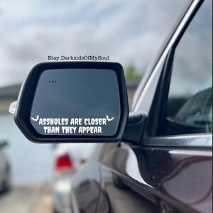 May include: A car side mirror with a white decal that reads "ASS HOLES ARE CLOSER THAN THEY APPEAR" in a bold, sans-serif font. The decal has two bat silhouettes on either side of the text. The car is dark red.