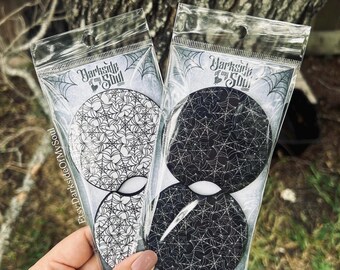 May include: Two packages of car coasters with a spiderweb design. The coasters are black and white, with the text "Darkside of my Soul" printed on the packaging. The coasters are packaged in clear plastic.