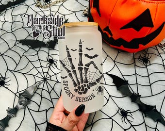 May include: A white frosted glass with a black and white design of a skeleton hand making a peace sign with bats and spiders. The text "Spooky Season" is written on the glass.