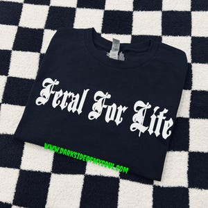 Pode incluir: Camiseta preta com o texto branco "Feral For Life" em fonte g&oacute;tica. A camisa &eacute; exibida em uma superf&iacute;cie xadrez em preto e branco. O endere&ccedil;o do site www.darksideofmysoul.com &eacute; impresso em verde na parte inferior.