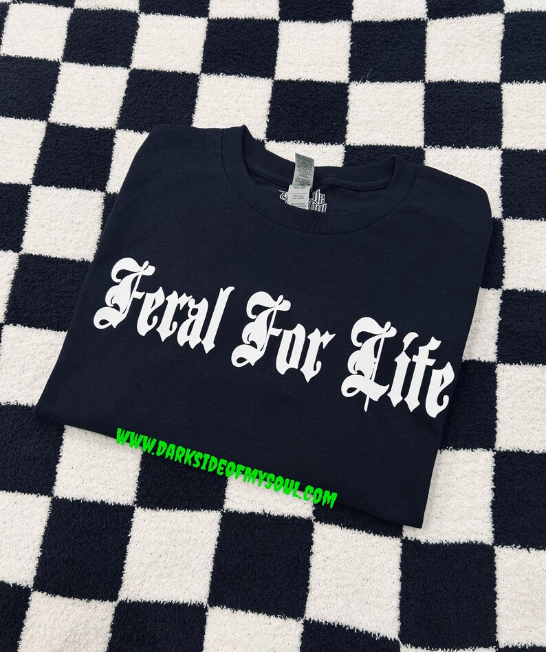 Pode incluir: Camiseta preta com o texto branco "Feral For Life" em fonte g&oacute;tica. A camisa &eacute; exibida em uma superf&iacute;cie xadrez em preto e branco. O endere&ccedil;o do site www.darksideofmysoul.com &eacute; impresso em verde na parte inferior.