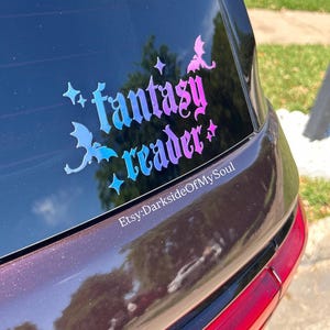 May include: A holographic sticker on a car window with the words "fantasy reader" in a stylized font. The sticker features stars and dragon silhouettes, with a color gradient from blue to pink. The Etsy shop name is visible at the bottom.