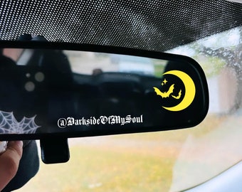 May include: A rearview mirror with a yellow crescent moon and two bats sticker. The sticker also includes two small stars. The text "@DarksideOfMySoul" is visible in the mirror.