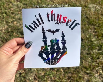 May include: A square white decal featuring the black text "hail thyself" in a gothic font, with a small black bat silhouette above a skeletal hand making a rock-and-roll gesture. The design has a holographic finish that reflects light.
