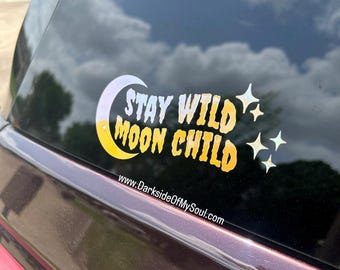 May include: A car window with a gold and white vinyl sticker that says "Stay Wild Moon Child" with a crescent moon and stars. The website www.DarksideOfMySoul.com is printed below the sticker.