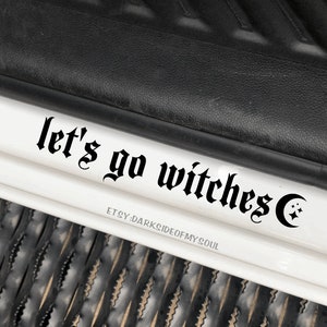 May include: Black and white decal with the text "let's go witches" in a gothic font, with a crescent moon and stars to the right of the text.