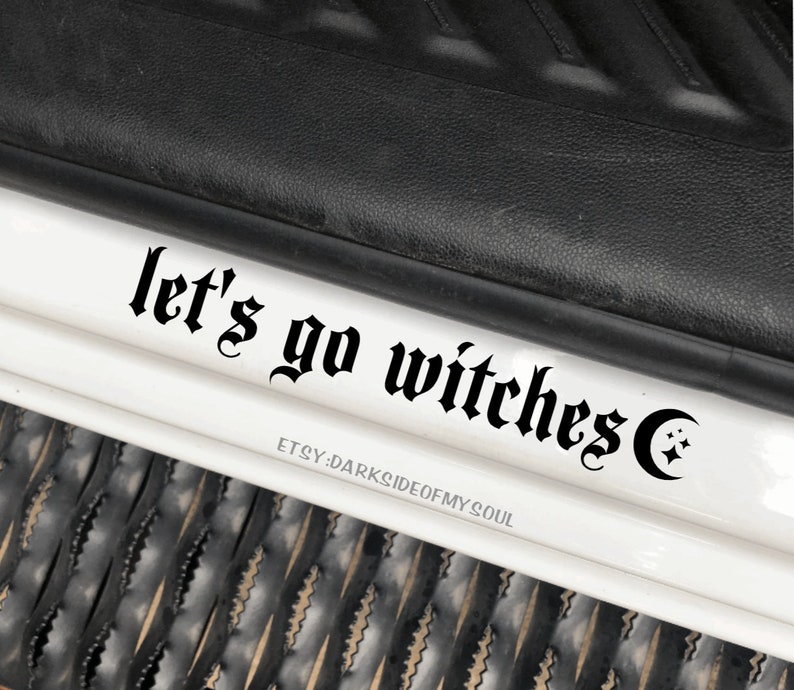 May include: Black and white decal with the text "let's go witches" in a gothic font, with a crescent moon and stars to the right of the text.