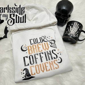 May include: White hooded sweatshirt with the text "Cold Brew Coffins Covens" in black and gold lettering. The design also includes a crescent moon, stars, and a witch's hat.