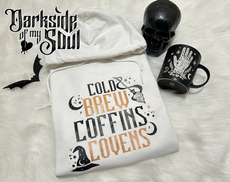 May include: White hooded sweatshirt with the text "Cold Brew Coffins Covens" in black and gold lettering. The design also includes a crescent moon, stars, and a witch's hat.