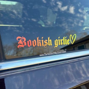 May include: A car window with a rainbow gradient vinyl sticker that says "Bookish girlie" with a heart.