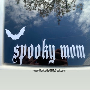 May include: A white vinyl decal with a bat and the words "spooky mom" in a gothic font. The decal is on a black car window. The website www.DarksideOfMySoul.com is visible at the bottom of the image.