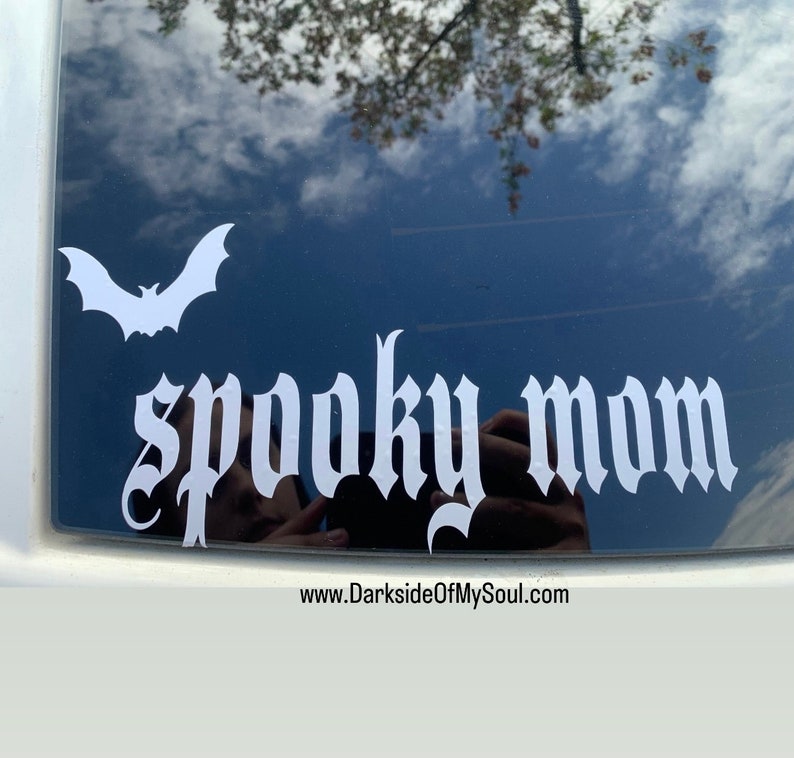 May include: A white vinyl decal with a bat and the words "spooky mom" in a gothic font. The decal is on a black car window. The website www.DarksideOfMySoul.com is visible at the bottom of the image.