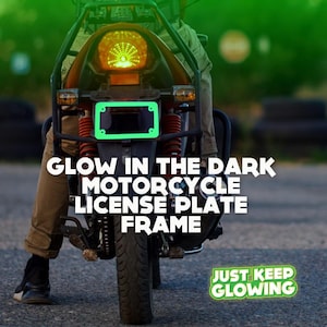 May include: A green glow-in-the-dark motorcycle license plate frame. The frame is attached to a motorcycle with a rider sitting on it. The text "JUST KEEP GLOWING" is visible in the lower right corner of the image.