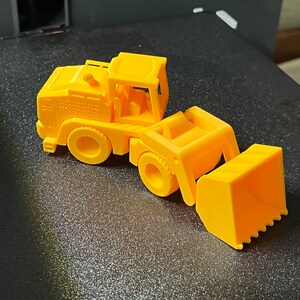 May include: A bright yellow toy front-end loader, featuring a detailed cab, wheels, and a functional-looking bucket. The toy is made of plastic and sits on a dark surface. The design is reminiscent of construction vehicles.